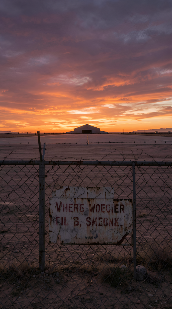 Art Style: Photorealistic Color Palette: orange, purple, pink, brown, gray Composition: Centered Content Type: Photograph Lighting Style: Natural Location Type: Outdoor Mood/Tone: Melancholic, Serene Perspective: Eye-level Primary Subjects: airstrip, hangar, chain-link fence, sign Time of Day: Sunset
